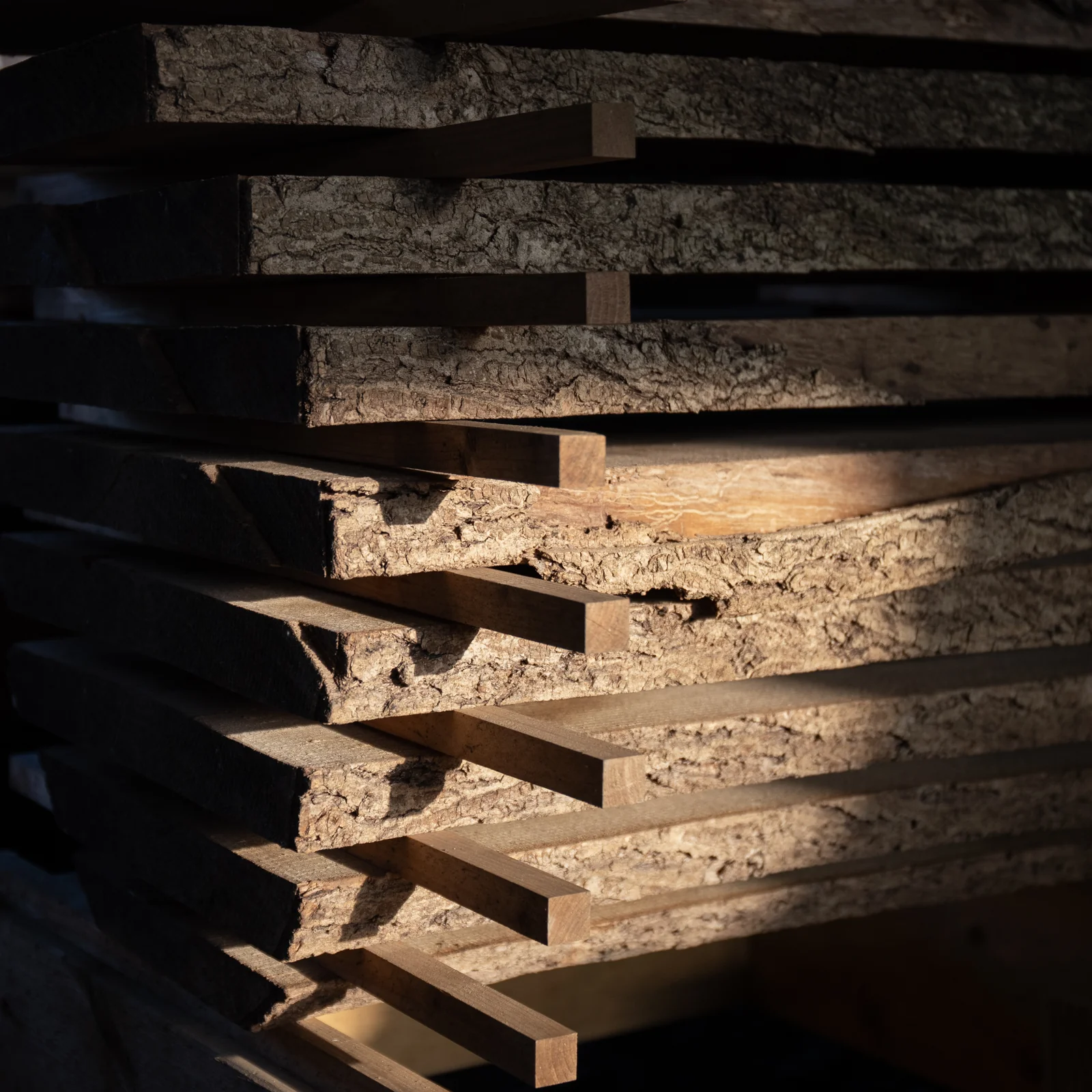 Stacked waney-edged hardwood boards with bark still on, interleaved with stickers for air drying, lit by raking light that casts sharp shadows across the grain